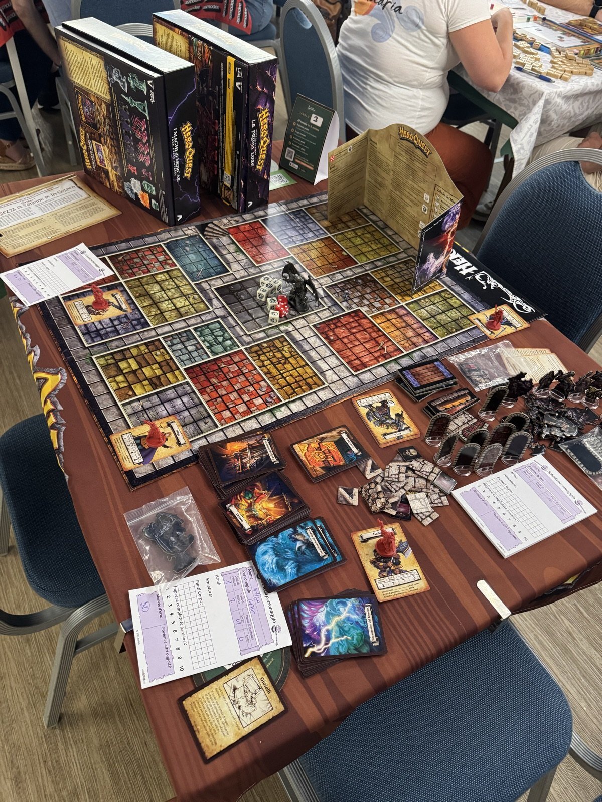 Un tavolo della DDCON con una partita a HeroQuest in corso: la plancia del dungeon, le miniature, i mazzi di carte e le schede personaggio compilate a mano, con il cartellino del tavolo e le scatole del gioco preso in ludoteca sullo sfondo