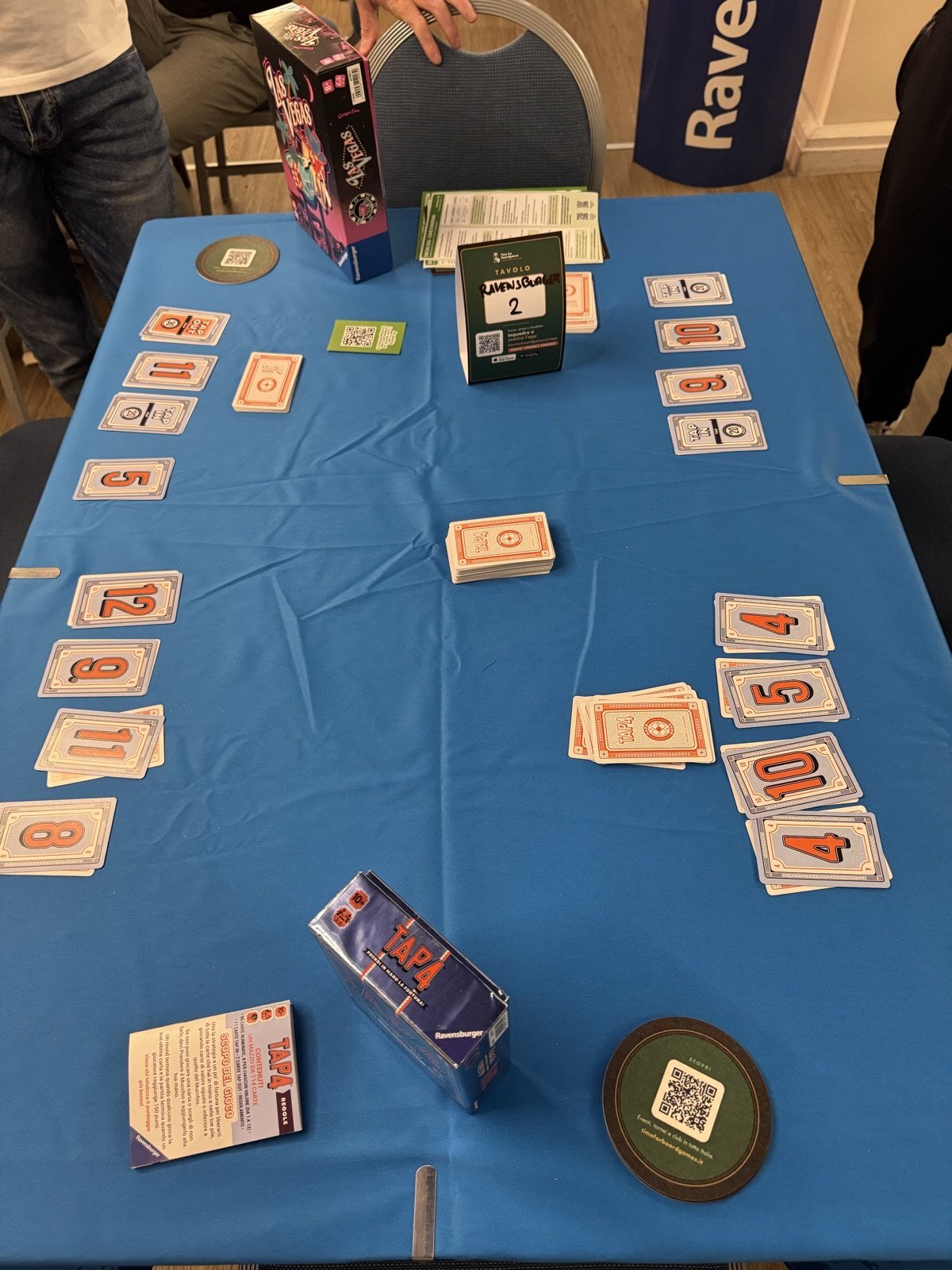 Un tavolo della DDCON col telo azzurro preparato per una partita a carte: al centro il cartellino di Time For Boardgames con scritto a pennarello «TAVOLO RAVENSBURGER 2» e il QR per prenotare, e in basso un sottobicchiere TfB. Sullo sfondo il banner Ravensburger