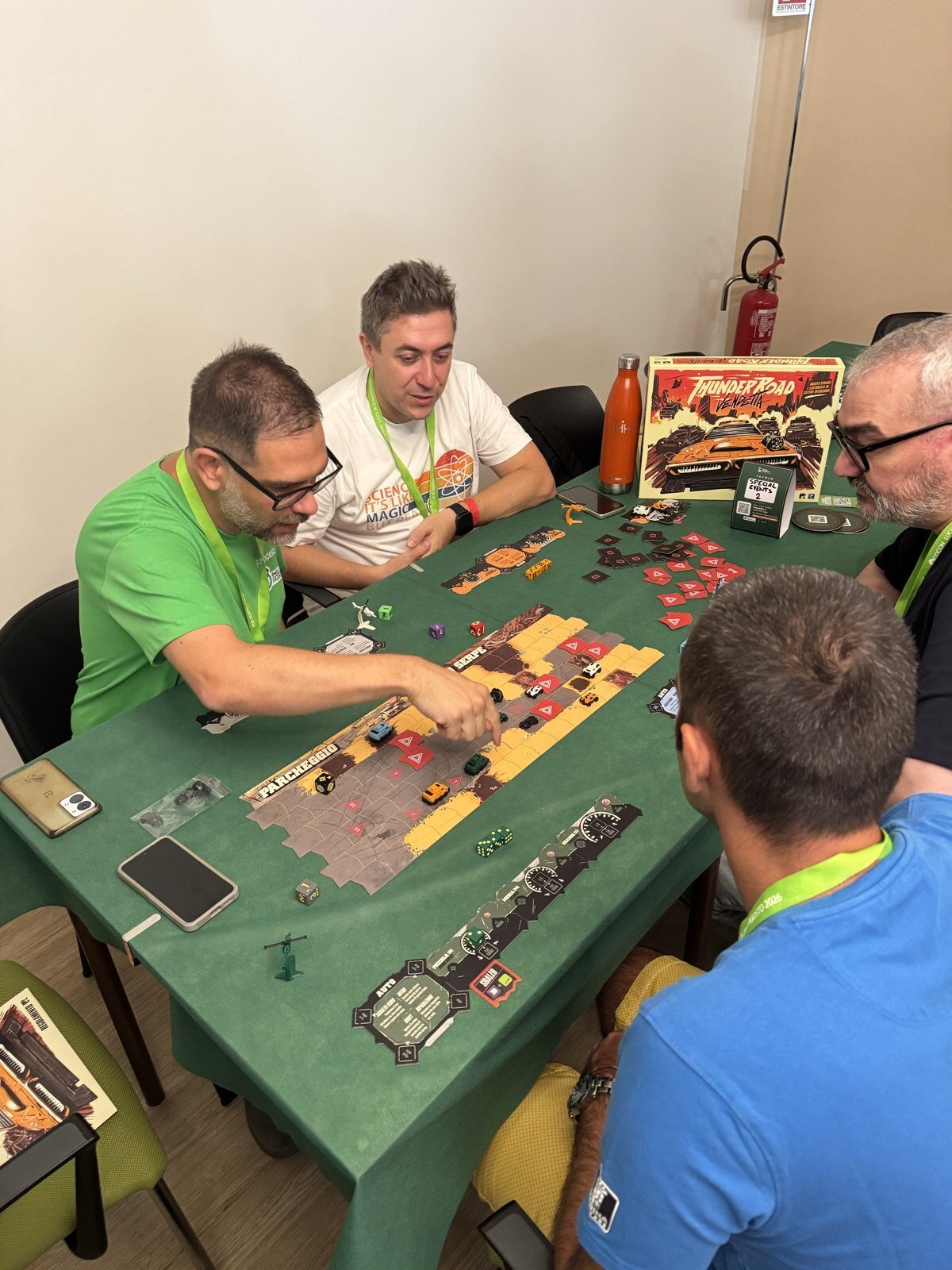 Quattro persone attorno a un tavolo dal telo verde alla DDCON, in piena partita a Thunder Road: Vendetta. Al centro del tavolo la plancia con le automobiline e i dadi; sul fondo la scatola del gioco e il cartellino di Time For Boardgames che segna il tavolo