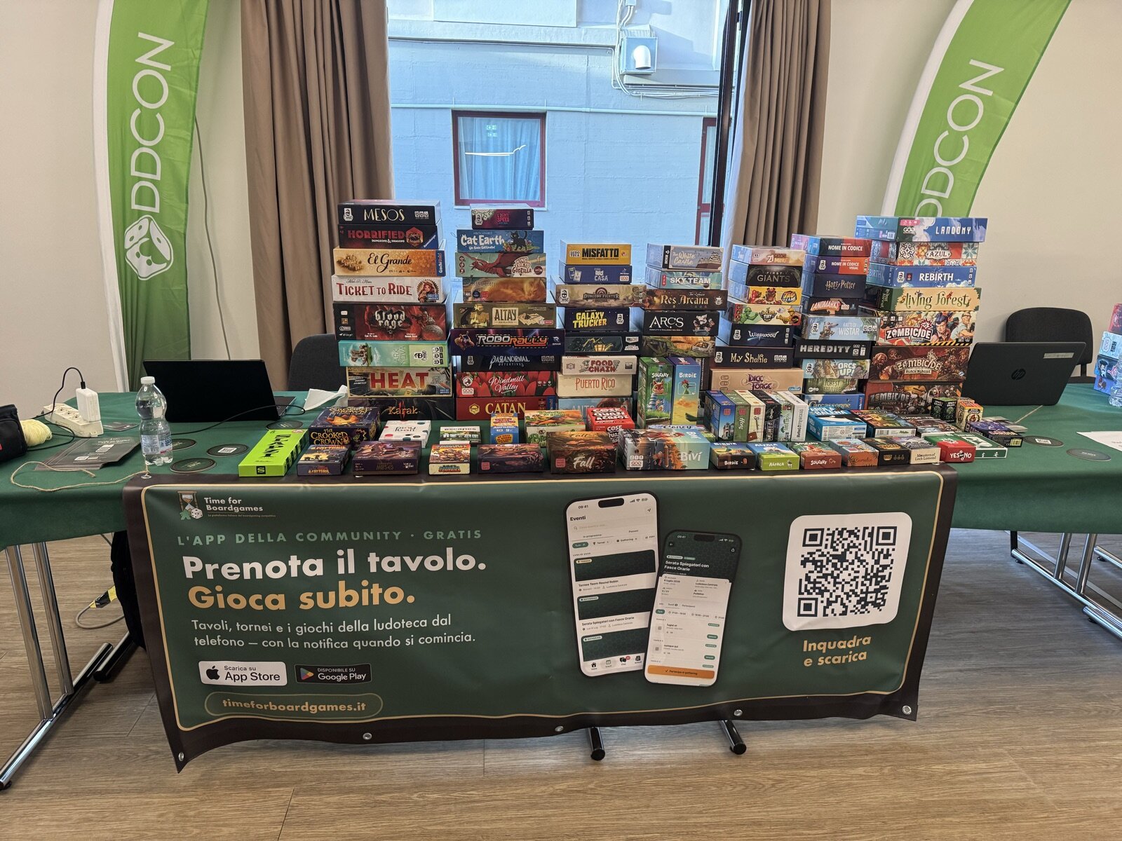 Il banco della ludoteca della DDCON: due file di scatole impilate su un tavolo con il telo verde, i roll-up DDCON alle spalle e sul davanti lo striscione di Time For Boardgames «Prenota il tavolo. Gioca subito.» con il QR per scaricare l'app